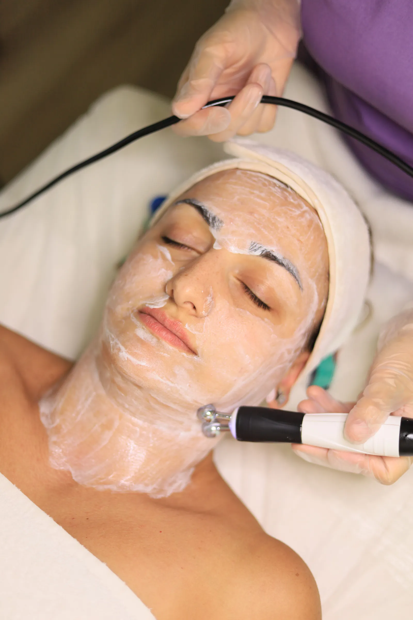 Client receiving a clinic facial treatment with a handheld skincare device while wearing a treatment mask.